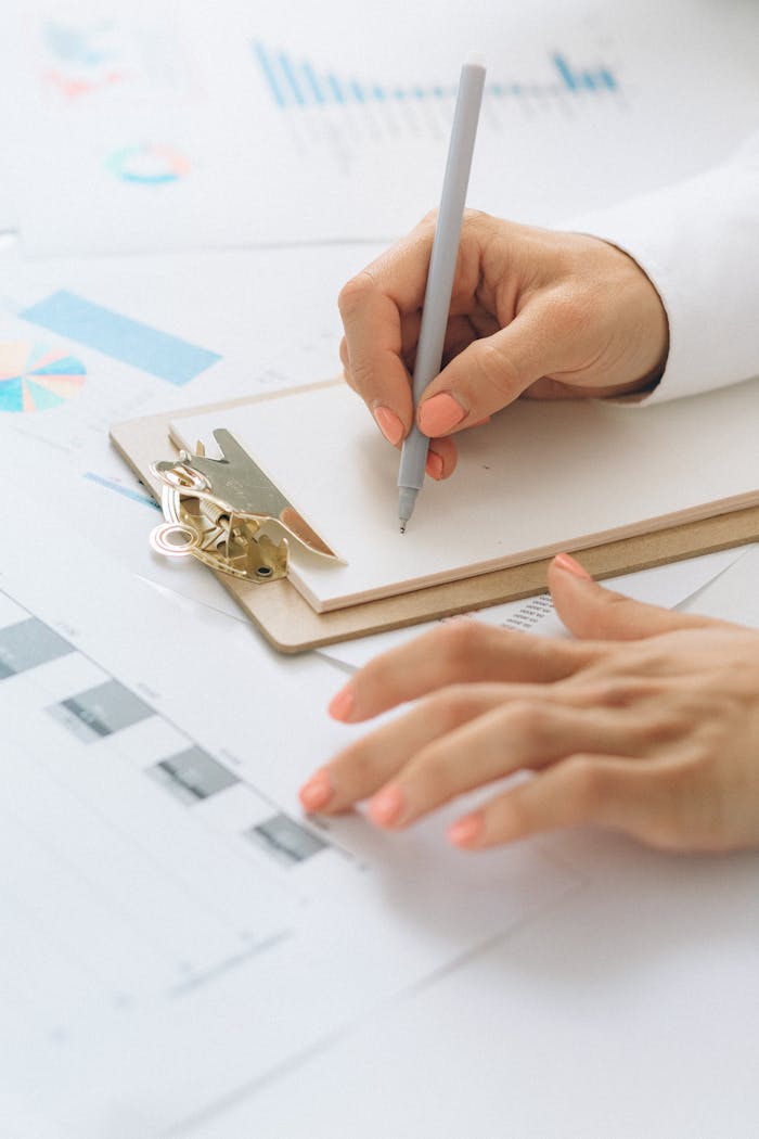 Detail of hands writing with a pen on a clipboard over financial papers.